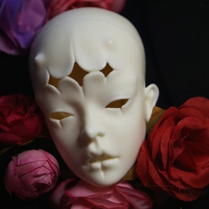 May include: A pale, blank doll head with cut-out shapes on the forehead and eyes. The doll head is surrounded by red and pink roses against a dark background. The head has a smooth, matte finish.