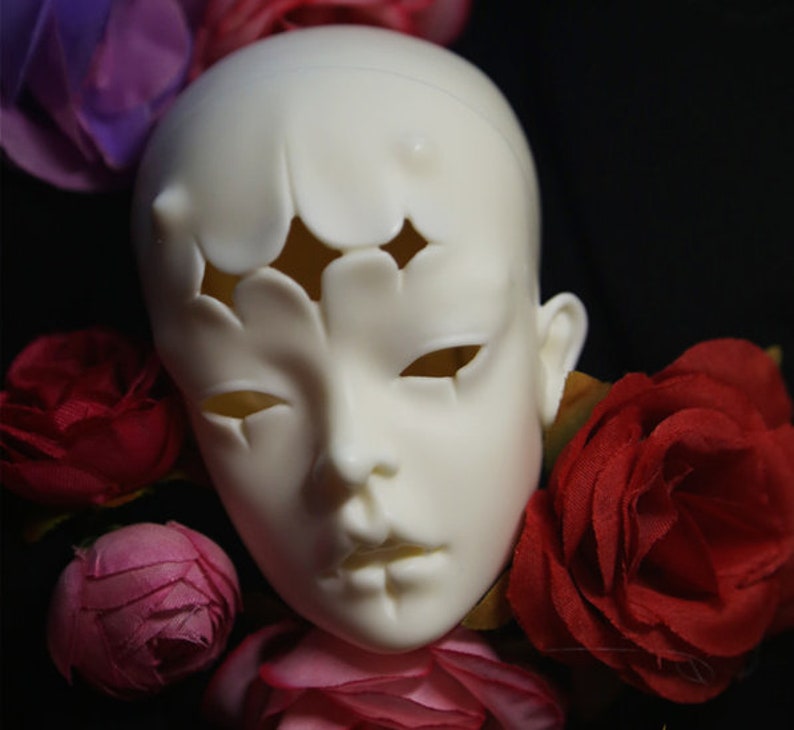 May include: A pale, blank doll head with cut-out shapes on the forehead and eyes. The doll head is surrounded by red and pink roses against a dark background. The head has a smooth, matte finish.