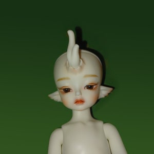 May include: A pale doll with a calm expression, featuring delicate facial details like subtle blush and defined eyes. The doll has a unique headpiece and pointed ears, set against a green backdrop. The doll's body is unadorned.