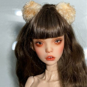 May include: A doll with brown hair and a fair complexion. The doll has a neutral expression with orange eye shadow and lipstick. The doll is wearing a headband with two fluffy, light brown pom-poms.