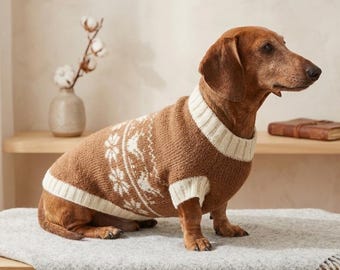 Può includere: Un bassotto marrone con un maglione marrone e bianco fantasia. Il maglione presenta un colletto, polsini e un motivo a fiocco di neve bianchi. Il cane è seduto su una coperta grigia su un tavolo di legno.