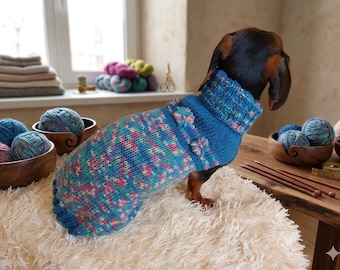 Può includere: Un bassotto indossa un maglione lavorato a mano blu, rosa e verde con collo alto e dettagli floreali. Il cane è su una superficie bianca e soffice, con filato e materiali per lavorare a maglia nelle vicinanze.