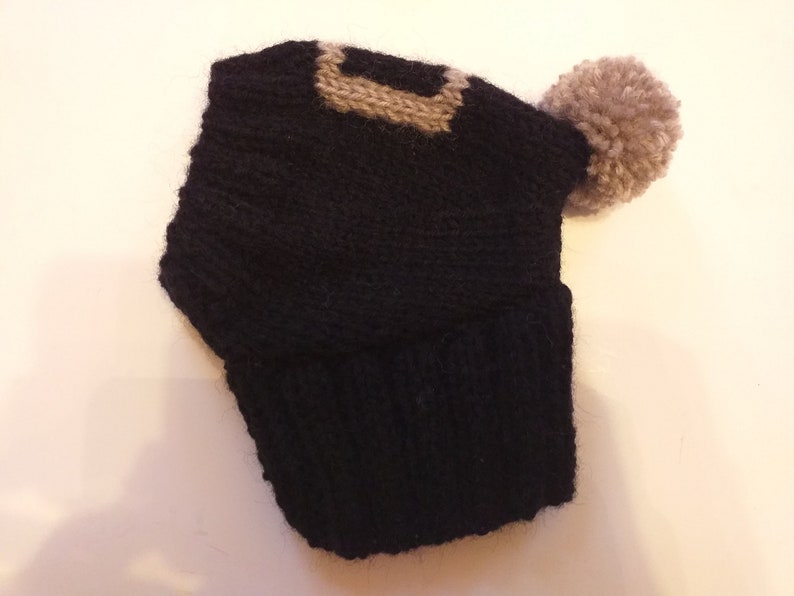 May include: A black knit hat with a tan geometric design on the front and a matching pom-pom on the side. The hat is made of a soft, textured yarn, perfect for cold weather.