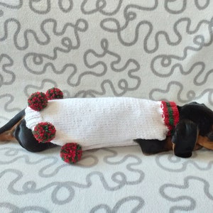 Puede incluir: Un perro salchicha con un su&eacute;ter blanco tejido con adornos rojos y verdes. El su&eacute;ter est&aacute; adornado con pompones rojos y verdes. El perro est&aacute; acostado sobre una superficie gris clara estampada.