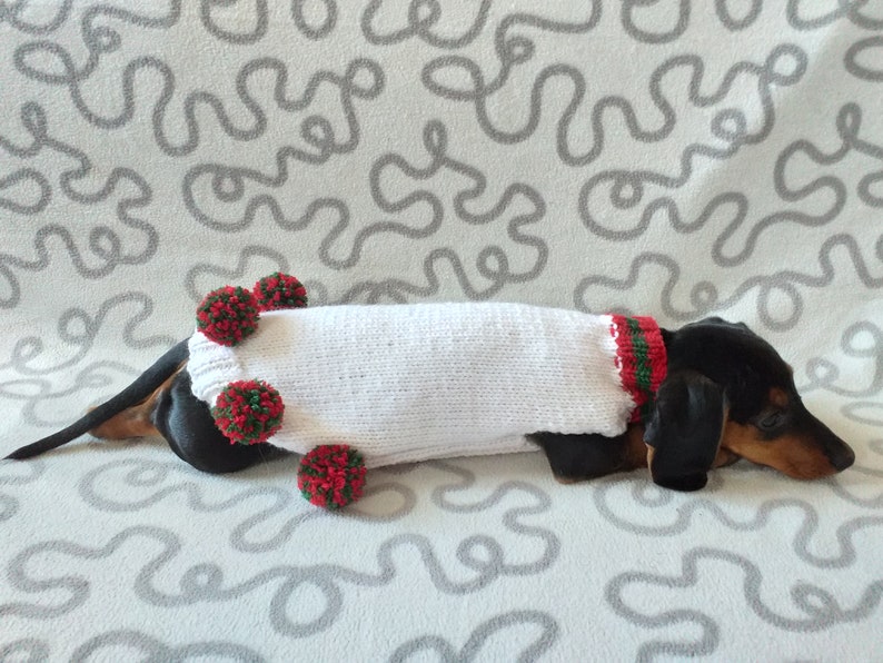 Puede incluir: Un perro salchicha con un su&eacute;ter blanco tejido con adornos rojos y verdes. El su&eacute;ter est&aacute; adornado con pompones rojos y verdes. El perro est&aacute; acostado sobre una superficie gris clara estampada.