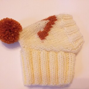 May include: A cream-colored knitted hat with a ribbed brim and a rust-colored pom-pom. The hat features a rust-colored design on the crown. The hat is made of soft yarn.