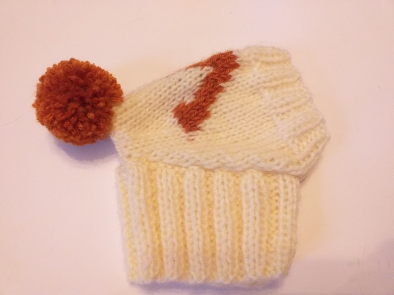 May include: A cream-colored knitted hat with a ribbed brim and a rust-colored pom-pom. The hat features a rust-colored design on the crown. The hat is made of soft yarn.