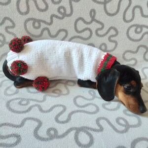 Puede incluir: Un perro salchicha con un su&eacute;ter blanco tejido con pompones y adornos rojos y verdes. El perro tiene pelaje negro y marr&oacute;n y est&aacute; acostado sobre una superficie estampada. El su&eacute;ter cubre el cuerpo del perro.