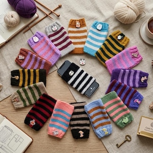 May include: A collection of knitted striped sleeves for mobile phones, each featuring a small decorative button. The sleeves are arranged in a circular pattern around a black smartphone. The knit covers display various color combinations and patterns.