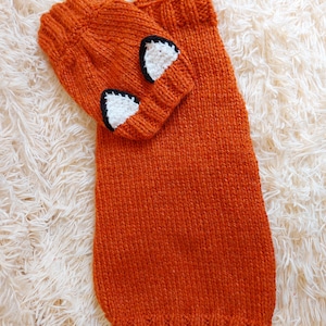 May include: An orange knitted hat and scarf set. The hat features white and black fox ear accents. The scarf is a long, rectangular shape. The set is made of a textured, knitted material.
