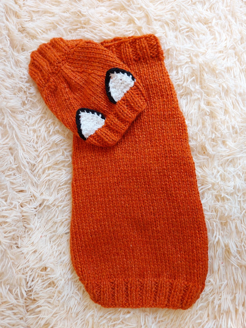 May include: An orange knitted hat and scarf set. The hat features white and black fox ear accents. The scarf is a long, rectangular shape. The set is made of a textured, knitted material.
