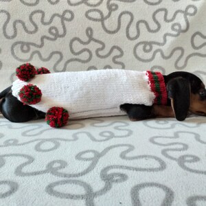 Puede incluir: Un perro salchicha negro y fuego con un su&eacute;ter blanco tejido con adornos rojos y verdes y pompones. El perro est&aacute; acostado sobre una superficie blanca estampada, mostrando un atuendo festivo para mascotas.