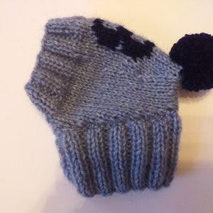 May include: A knitted beanie hat in shades of gray with a dark navy blue pom-pom and a matching design on the front. The hat has a ribbed brim and a soft, textured appearance, suitable for cold weather.