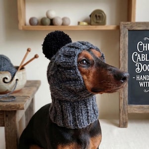 May include: A dachshund wearing a gray cable-knit dog hat with a black pom-pom. The hat covers the dog's head and neck. Text on a chalkboard reads "Chunky Cable-Knit Dog Hat - Handcrafted with Love".