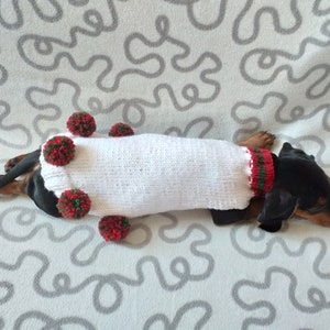 Puede incluir: Un perro salchicha negro y fuego con un su&eacute;ter blanco tejido con pompones rojos y verdes. El su&eacute;ter tiene un cuello y ribete a rayas rojas y verdes. El perro est&aacute; acostado sobre una superficie gris y blanca estampada.