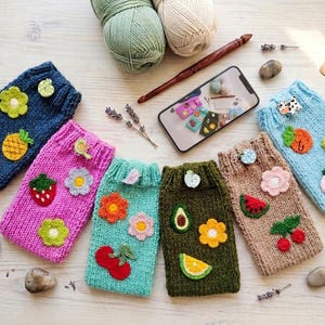 Peut inclure: Plusieurs étuis de téléphone tricotés à la main, de couleurs variées, ornés d'applications de fruits et de fleurs. Les étuis sont bleus, roses, verts et marron. Un crochet, de la laine et un smartphone affichant des étuis similaires sont également visibles.