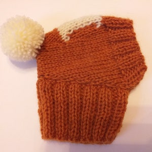 May include: A knitted hat in shades of orange and white, featuring a large cream-colored pom-pom. The hat has a ribbed brim and a white accent on the crown. This handmade hat is perfect for cold weather.