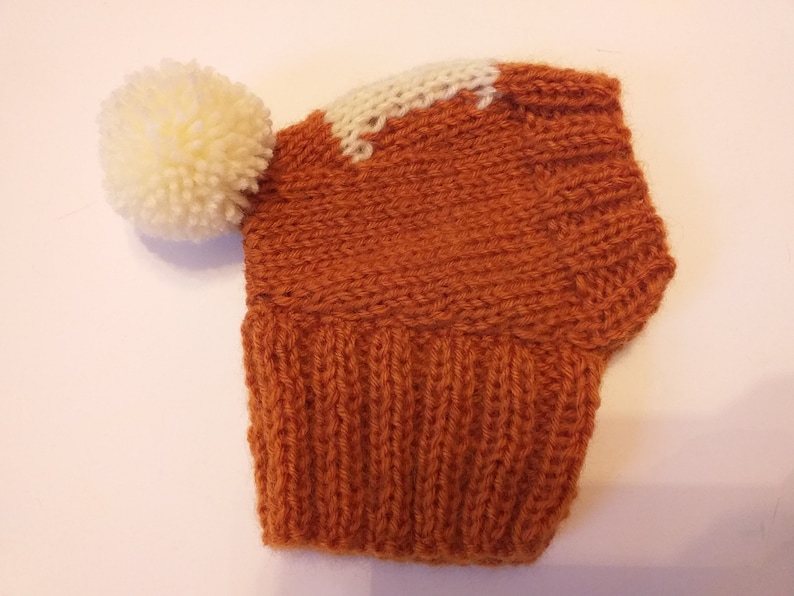 May include: A knitted hat in shades of orange and white, featuring a large cream-colored pom-pom. The hat has a ribbed brim and a white accent on the crown. This handmade hat is perfect for cold weather.