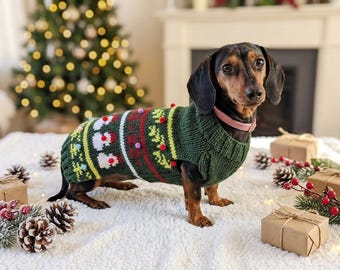 Può includere: Un cane bassotto indossa un maglione natalizio verde con un motivo festivo di pupazzi di neve, alberi e pompon colorati. Il cane è in piedi su una superficie bianca, circondato da regali confezionati, pigne e decorazioni natalizie.