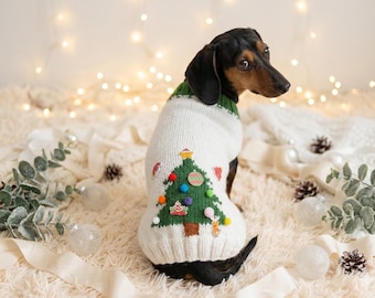 Può includere: Un cane bassotto indossa un maglione natalizio festivo. Il maglione bianco presenta un design ad albero di Natale verde con ornamenti colorati e un colletto verde. Il cane è seduto su una superficie bianca e soffice con decorazioni natalizie.