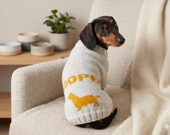 May include: A light gray knit dog sweater featuring a mustard yellow graphic of a dachshund silhouette and the text COOPER in a matching mustard yellow font. The sweater has a ribbed collar and hem, designed for a small canine with short fur.
