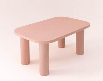 May include: A small, rectangular table in a soft pink hue. The table has rounded corners and four cylindrical legs. The design is minimalist and modern, suitable for various interior styles. The table's dimensions are approximately 24 inches long, 16 inches wide, and 12 inches high.