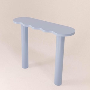 Puede incluir: Una consola azul claro con un borde ondulado. La mesa tiene una parte superior rectangular y tres patas cilíndricas. La mesa está pintada en un color azul pastel sólido y está colocada sobre un fondo neutro.
