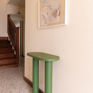 以下が含まれることがあります： 2本の円筒形の脚が付いた、緑色の半円形のコンソールテーブル。テーブルはクリーム色の壁に立てかけられ、上には額入りのアートワークがあります。床はテラゾー模様です。背景には階段が見えます。