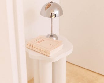 May include: A small, white, three-legged side table supports a silver-colored dome lamp and two books. The books have light-colored covers with text. The lamp is on a white table in a room with a light-colored wall.