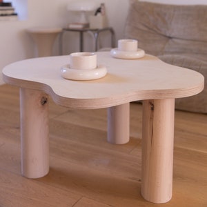 May include: A light wood coffee table with a unique, organic shape and four cylindrical legs. Two white ceramic cups and saucers are placed on the table.