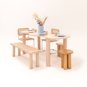 May include: A light wood dining table with a wavy edge and four chairs. The table is set with plates and cups. A light wood bench is in front of the table. A light wood stool is in the foreground. A vase with dried flowers is on the table.