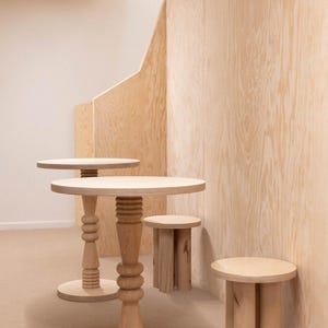 Może przedstawiać: Kolekcja jasnych drewnianych stoł&oacute;w i stołk&oacute;w. Dwa stoły mają okrągłe blaty i ozdobne, toczone nogi. Widoczne są r&oacute;wnież dwa mniejsze stołki. Meble ustawione są na tle jasnej drewnianej ściany.