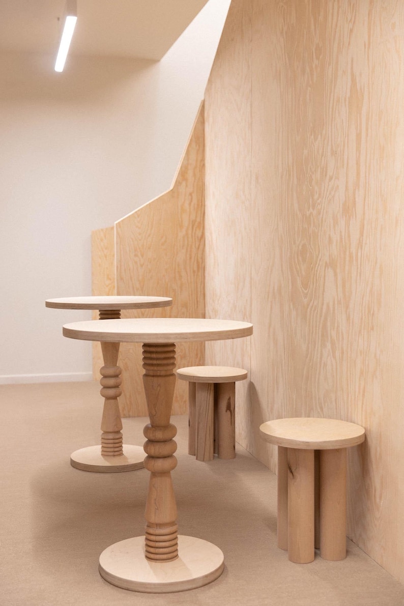 Może przedstawiać: Kolekcja jasnych drewnianych stoł&oacute;w i stołk&oacute;w. Dwa stoły mają okrągłe blaty i ozdobne, toczone nogi. Widoczne są r&oacute;wnież dwa mniejsze stołki. Meble ustawione są na tle jasnej drewnianej ściany.