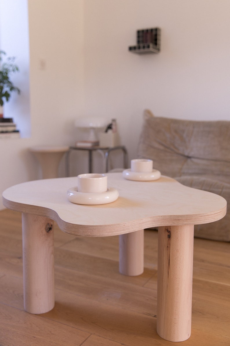 May include: A light wood coffee table with a unique, organic shape and four cylindrical legs. Two white ceramic cups and saucers are placed on the table.