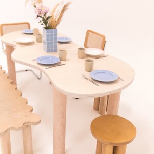 May include: A light wood dining table with a unique, organic shape and four legs. The table is set with blue and white plates, beige cups, and silverware. A small, light wood stool with four legs is in the foreground.
