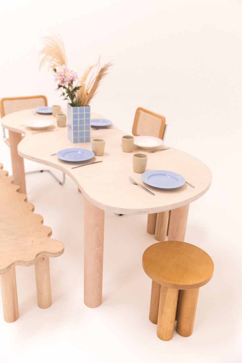 May include: A light wood dining table with a unique, organic shape and four legs. The table is set with blue and white plates, beige cups, and silverware. A small, light wood stool with four legs is in the foreground.