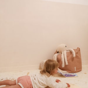 May include: A young child in a pink and white outfit is lying on a wooden balance board. The child is looking at a book on the floor. There is a brown storage bin with a white octopus toy in the background.