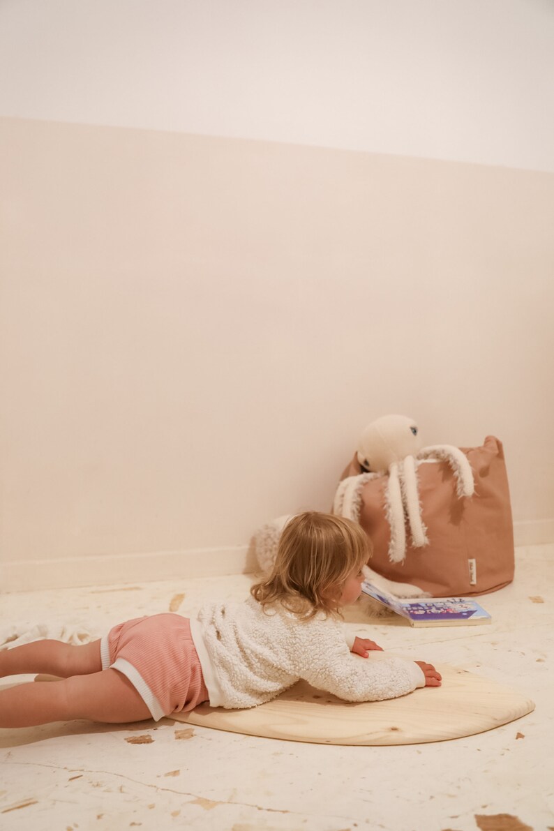May include: A young child in a pink and white outfit is lying on a wooden balance board. The child is looking at a book on the floor. There is a brown storage bin with a white octopus toy in the background.