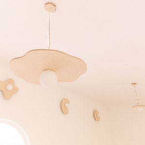 Pu&ograve; includere: Un set di cinque luci a sospensione in legno a forma di nuvole e uova. Le luci sono appese al soffitto e hanno lampadine bianche. Le luci sono di colore marrone chiaro.