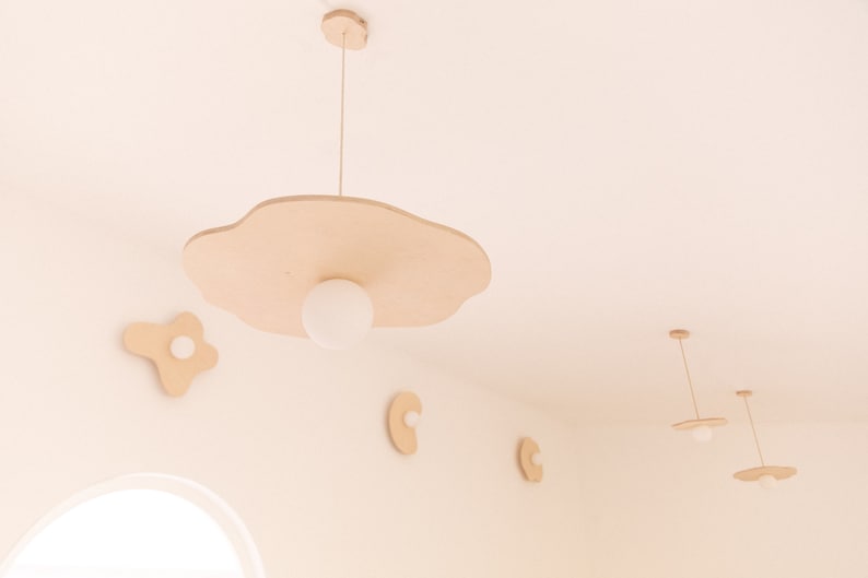 Pu&ograve; includere: Un set di cinque luci a sospensione in legno a forma di nuvole e uova. Le luci sono appese al soffitto e hanno lampadine bianche. Le luci sono di colore marrone chiaro.