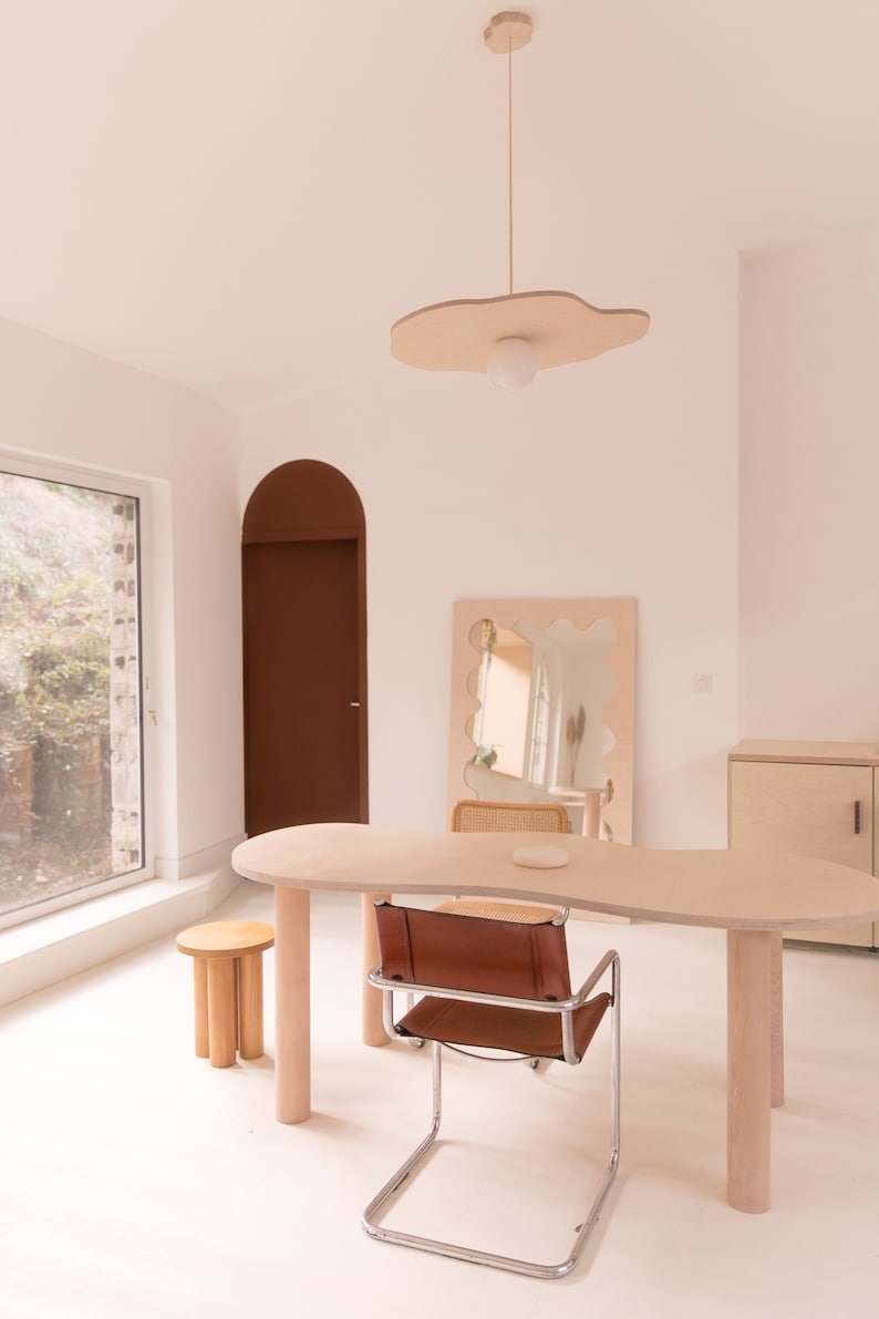 Puede incluir: Una mesa de madera clara con una forma org&aacute;nica y una silla de cuero marr&oacute;n. La mesa tiene una superficie blanca y est&aacute; sostenida por cuatro patas de madera. La silla tiene un marco de cromo y est&aacute; colocada a la derecha de la mesa. Un peque&ntilde;o taburete de madera est&aacute; en primer plano. La habitaci&oacute;n tiene paredes blancas y una gran ventana con vista a los &aacute;rboles.