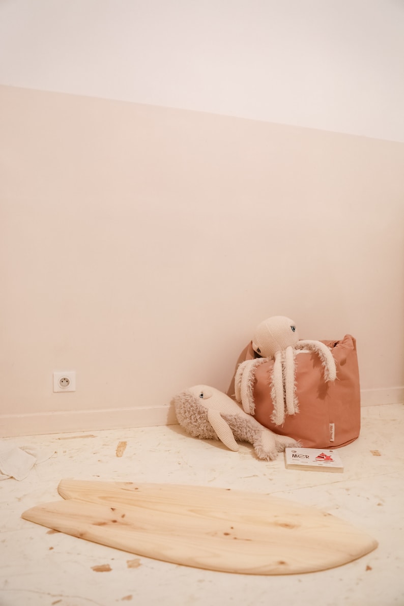 May include: A wooden surfboard-shaped balance board with a light brown finish. The board is on a light beige floor with a stuffed octopus and whale toy in the background.
