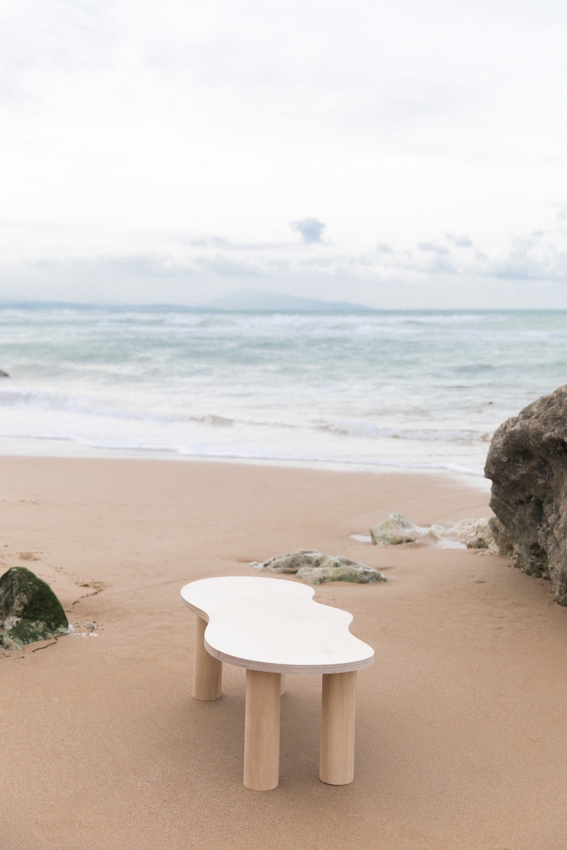 May include: White organic shaped coffee table with four wooden legs on a sandy beach.