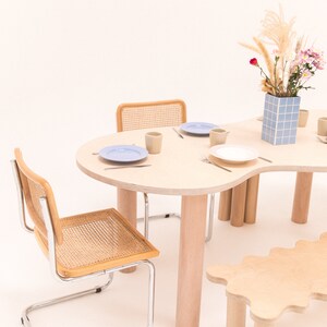 May include: A light wood dining table with a unique organic shape and four legs. Two chairs with woven cane seats and chrome frames are positioned around the table. A small light wood bench with a wavy edge and four legs is in the foreground. A vase with a blue and white tiled pattern holds a bouquet of dried grasses and pink flowers.