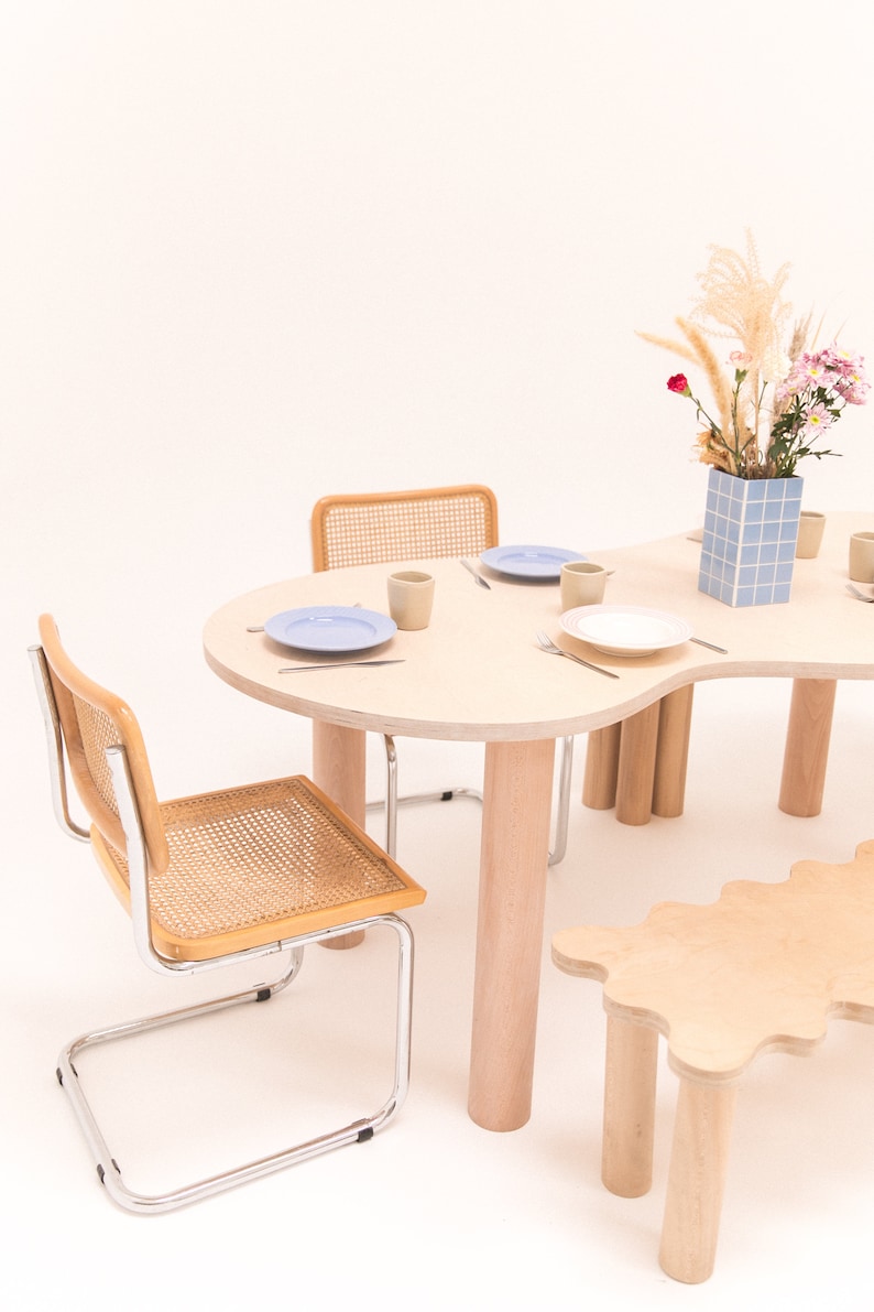 May include: A light wood dining table with a unique organic shape and four legs. Two chairs with woven cane seats and chrome frames are positioned around the table. A small light wood bench with a wavy edge and four legs is in the foreground. A vase with a blue and white tiled pattern holds a bouquet of dried grasses and pink flowers.