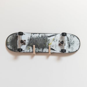 Puede incluir: Una tabla de skate negra y blanca con pintura desgastada est&aacute; montada en una pared blanca usando dos clavijas de madera.