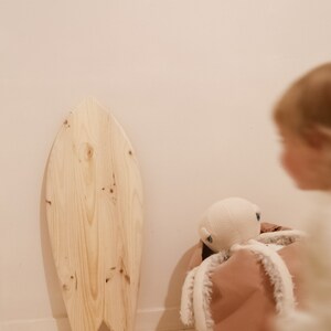 May include: A wooden surfboard toy with a rounded shape and a natural wood finish. The surfboard is leaning against a wall and there is a stuffed octopus toy in a pink storage bin in the foreground.