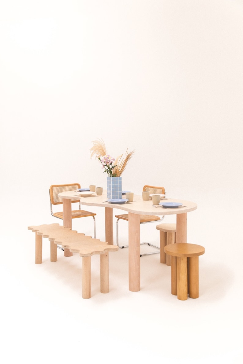 May include: A light wood dining table with a wavy edge and four chairs. The table is set with plates and cups. A light wood bench is in front of the table. A light wood stool is in the foreground. A vase with dried flowers is on the table.