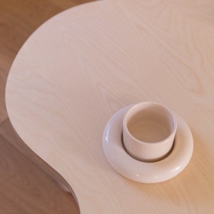 May include: A white ceramic cup and saucer set on a light wood table.