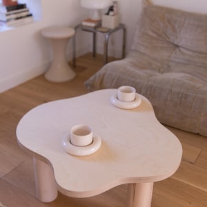 May include: A light wood coffee table with a unique cloud-shaped top and four rounded legs. Two white ceramic cups are placed on the table, each with a white saucer.